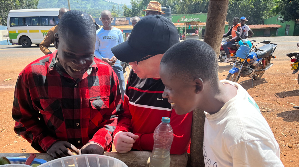 A man smiles alongside his wife and another man. There is a bus, motorbike and tree behind them.