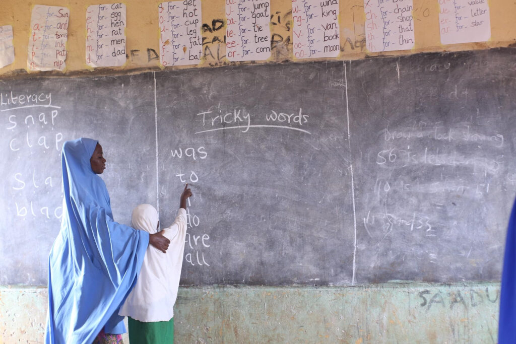 A female teacher is helping a girl to read words written in chalk on a blackboard.